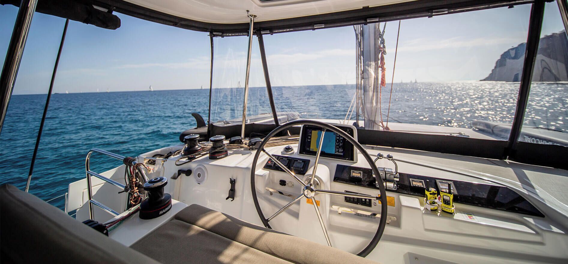 La cabina di pilotaggio di una barca con timone, pannelli di controllo e vista sul mare e su una scogliera.