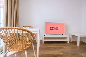 Ein Zimmer mit einem Korbstuhl, einem weißen Tisch, einem Fernseher, der 'GUEST READY' anzeigt, und einer weißen Bank.