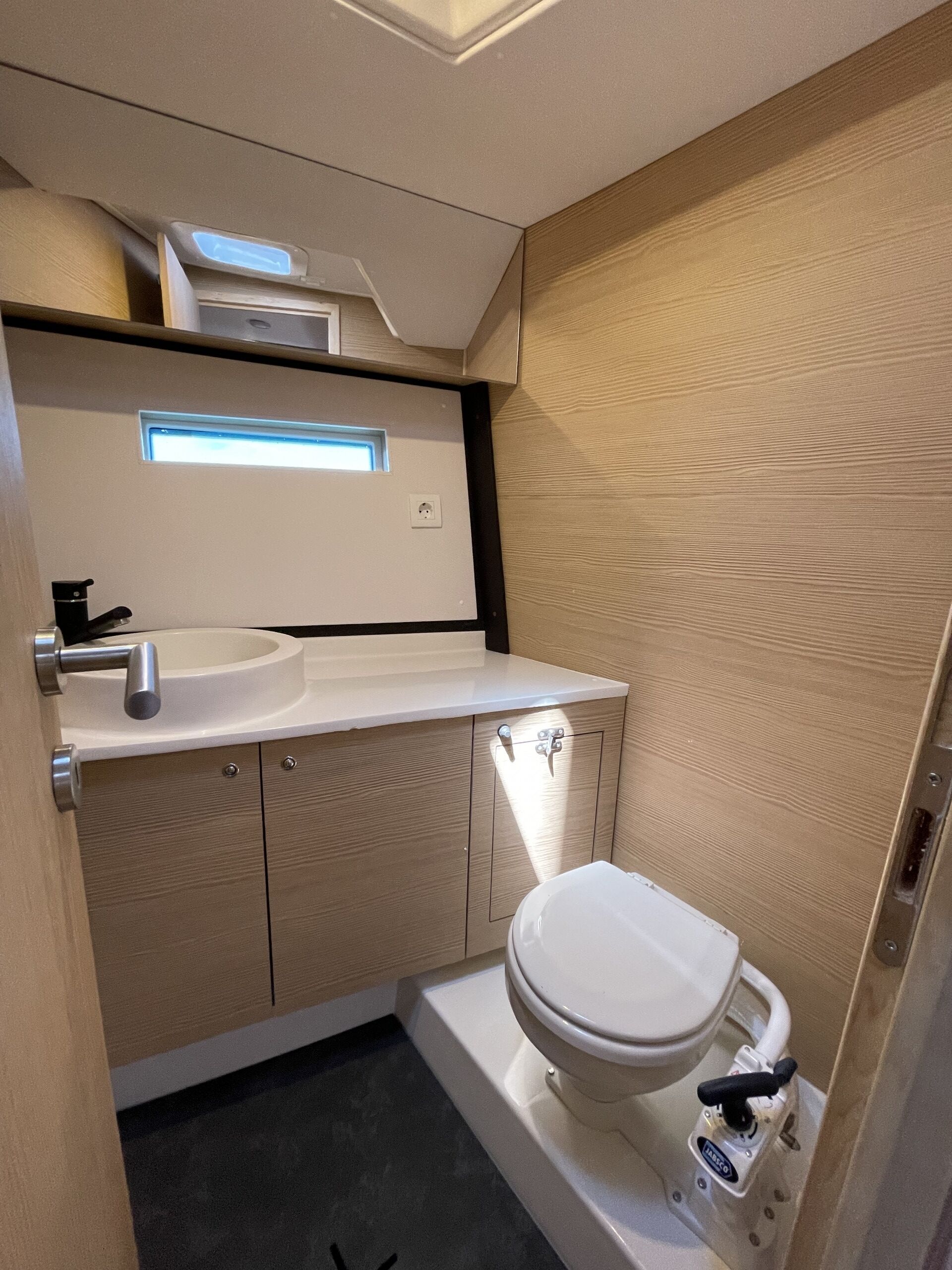 Une petite salle de bain avec un lavabo blanc, des toilettes et une petite fenêtre située au-dessus du lavabo.