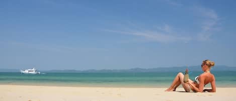 A beach with a boat in the water and a person sitting on the sand.