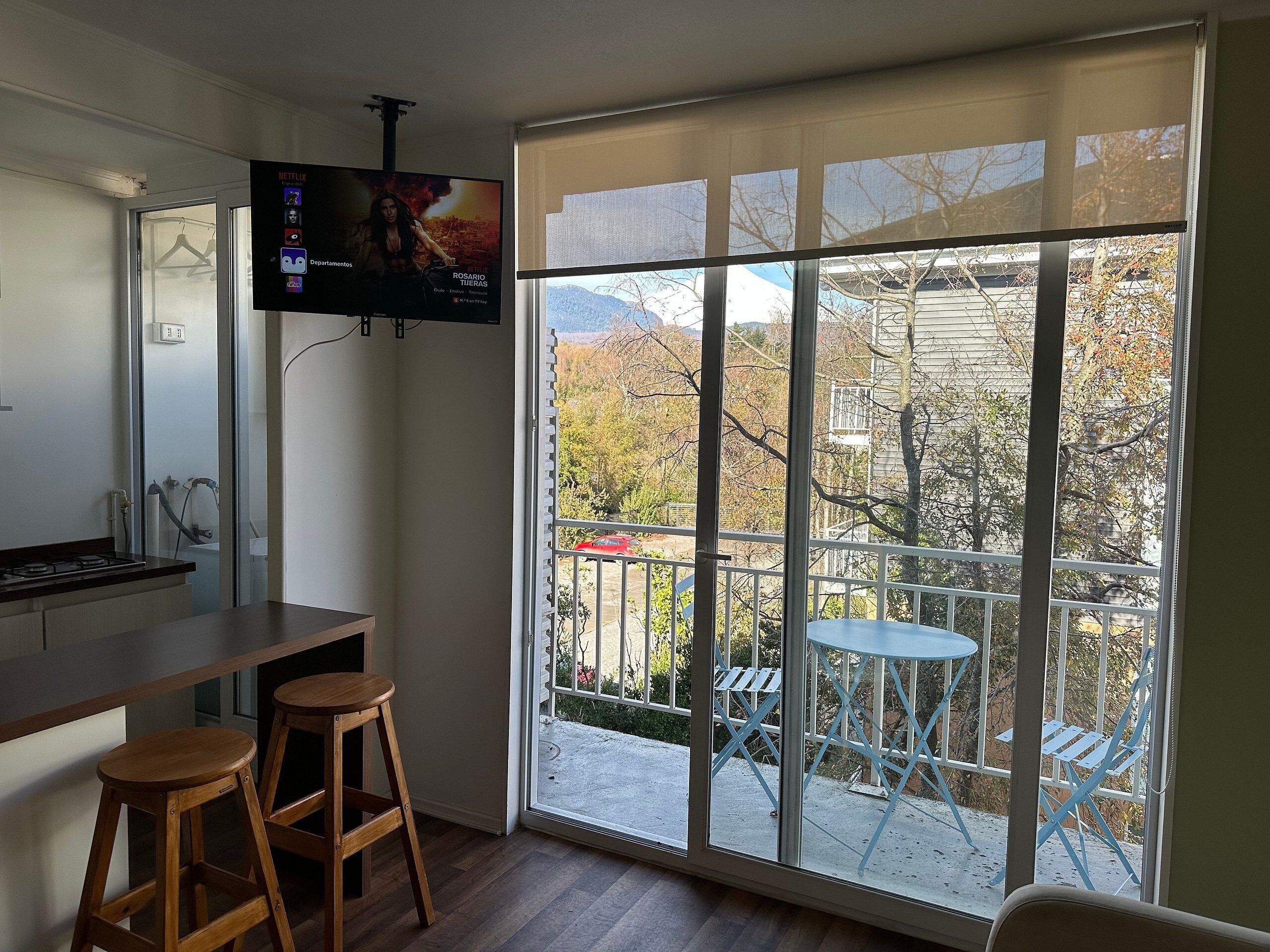 A room with a large window, a TV mounted on the wall, a kitchen area, and a balcony with outdoor furniture.