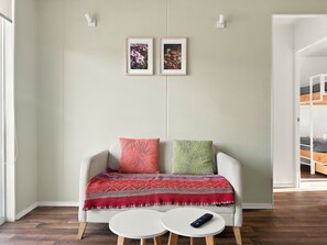 Una habitación con un sofá blanco, una manta estampada, un control remoto sobre una mesita y dos cuadros en la pared.