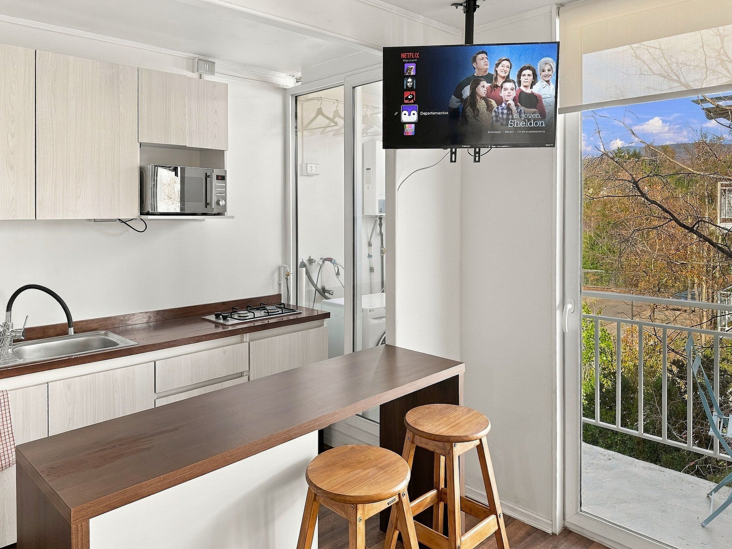 Una cocina moderna con un televisor de pantalla plana montado sobre la encimera, una estufa a gas y un lavabo. Hay dos taburetes de madera en la encimera.