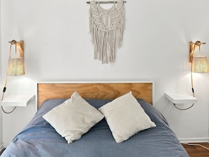 Quarto com cama, duas luminárias de cabeceira, um painel de parede em macramé e cabeceira de madeira.