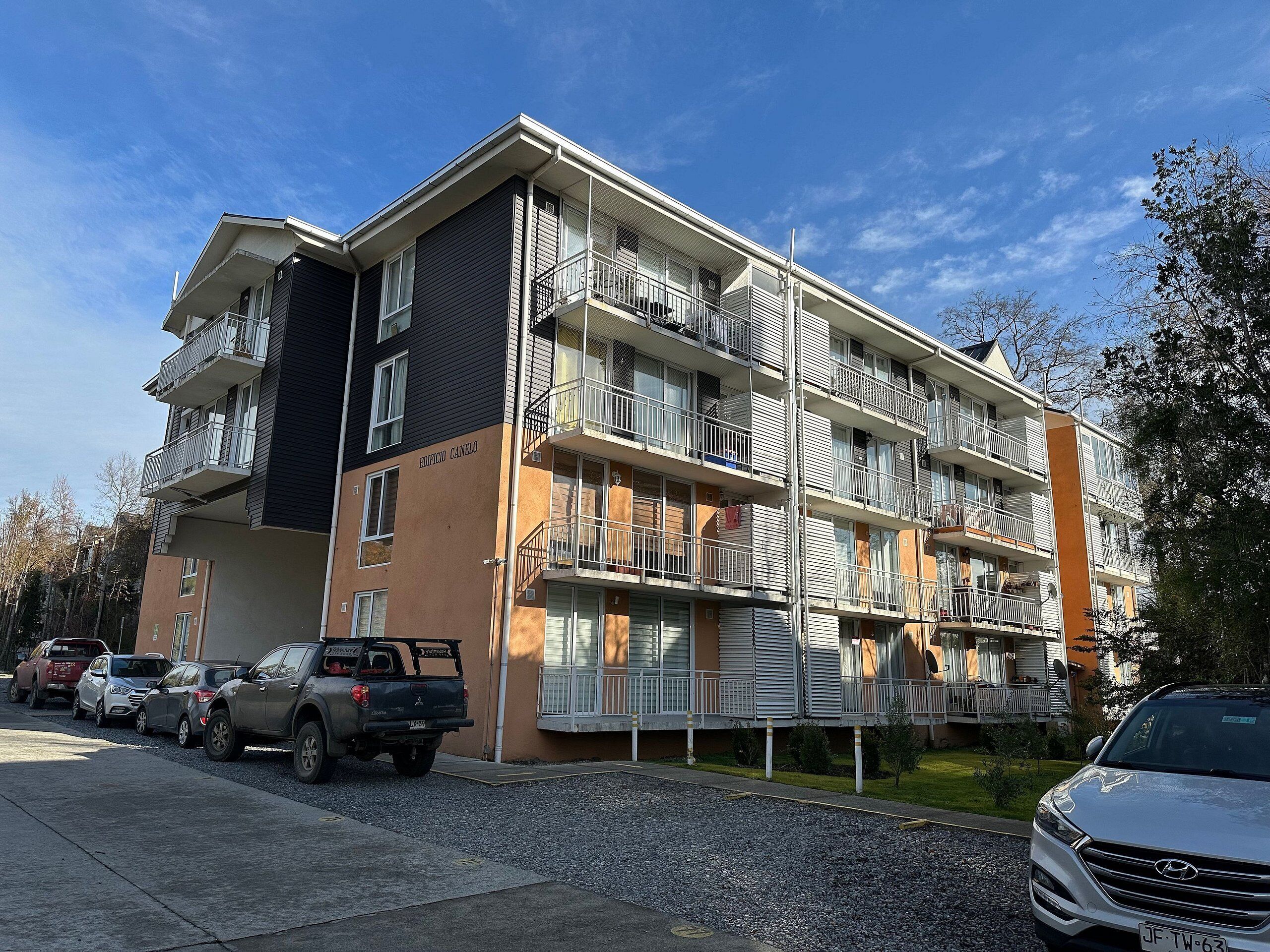 A multi-story apartment building with balconies, a parking area, and a clear sky.
