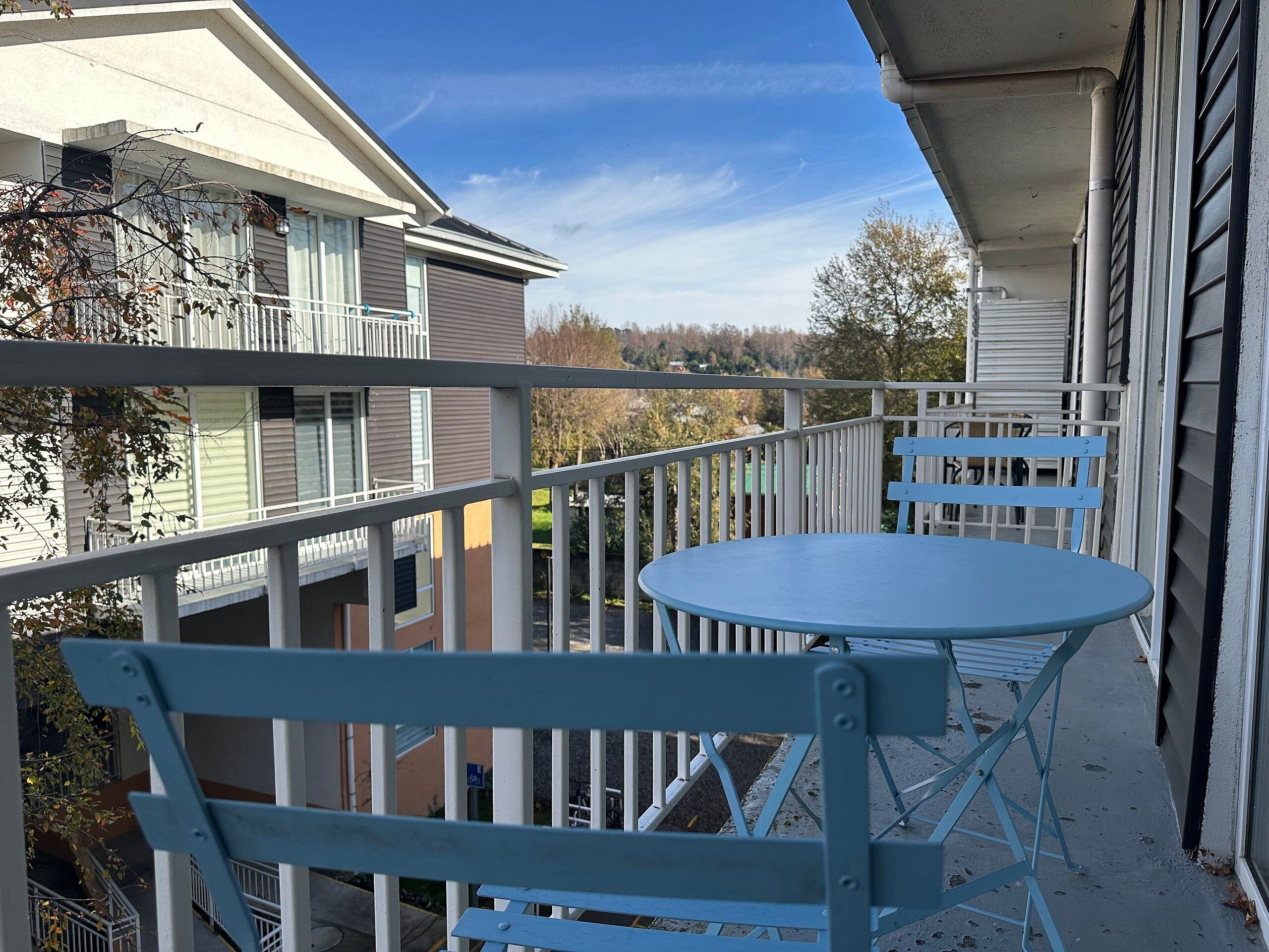 A balcony with a blue table and chairs, a white railing, and a view of neighboring buildings.