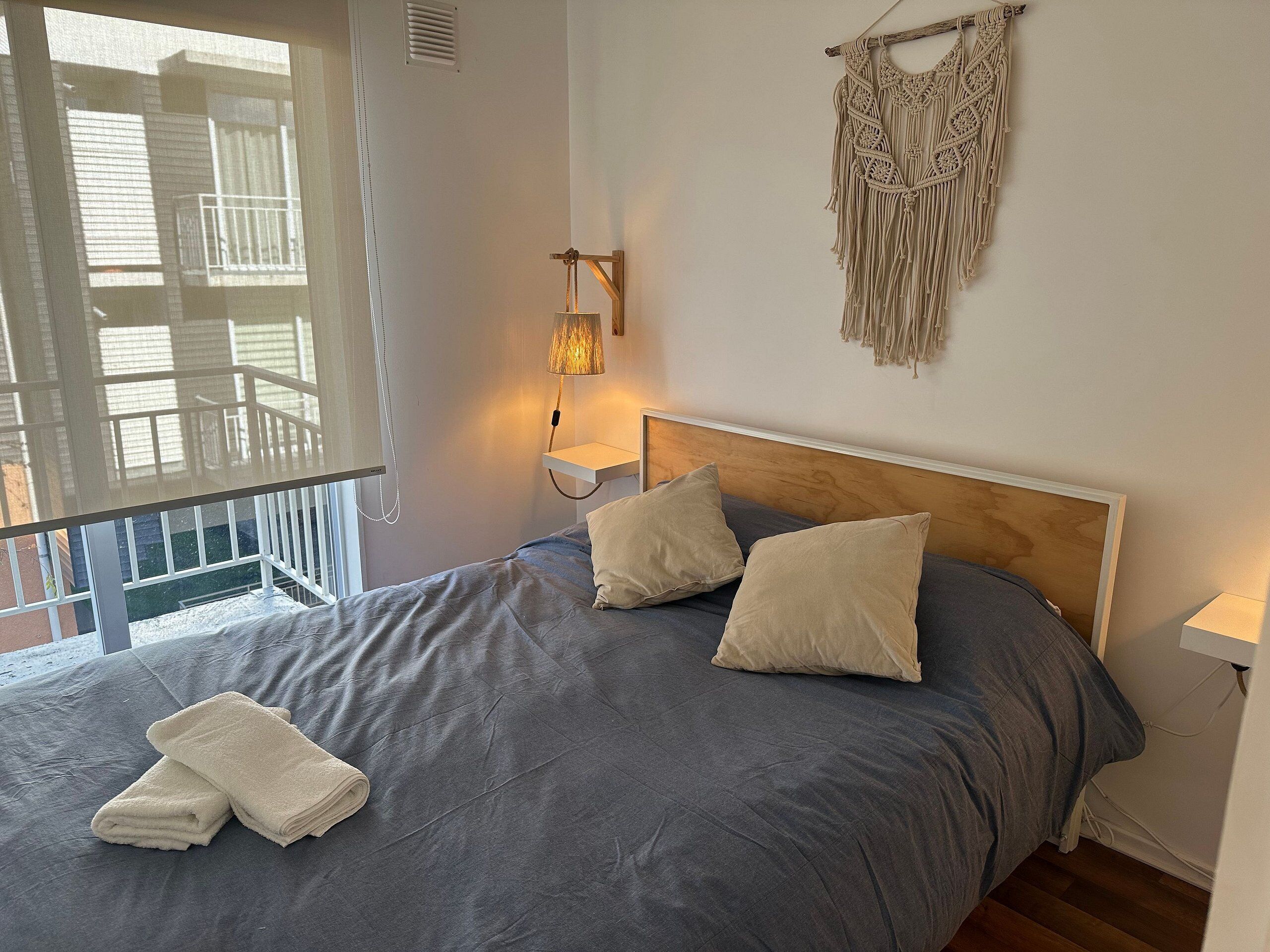 A bedroom with a bed, two pillows, a macrame wall hanging, a bedside lamp, and a window with a view of a balcony.