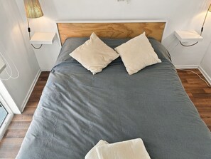 Un lit bien fait, recouvert d’une couvre-lit grise et agrémenté de deux oreillers beige, est placé de part et d’autre de tables de chevet sur lesquelles reposent des lampes, dans une pièce au plancher en bois.