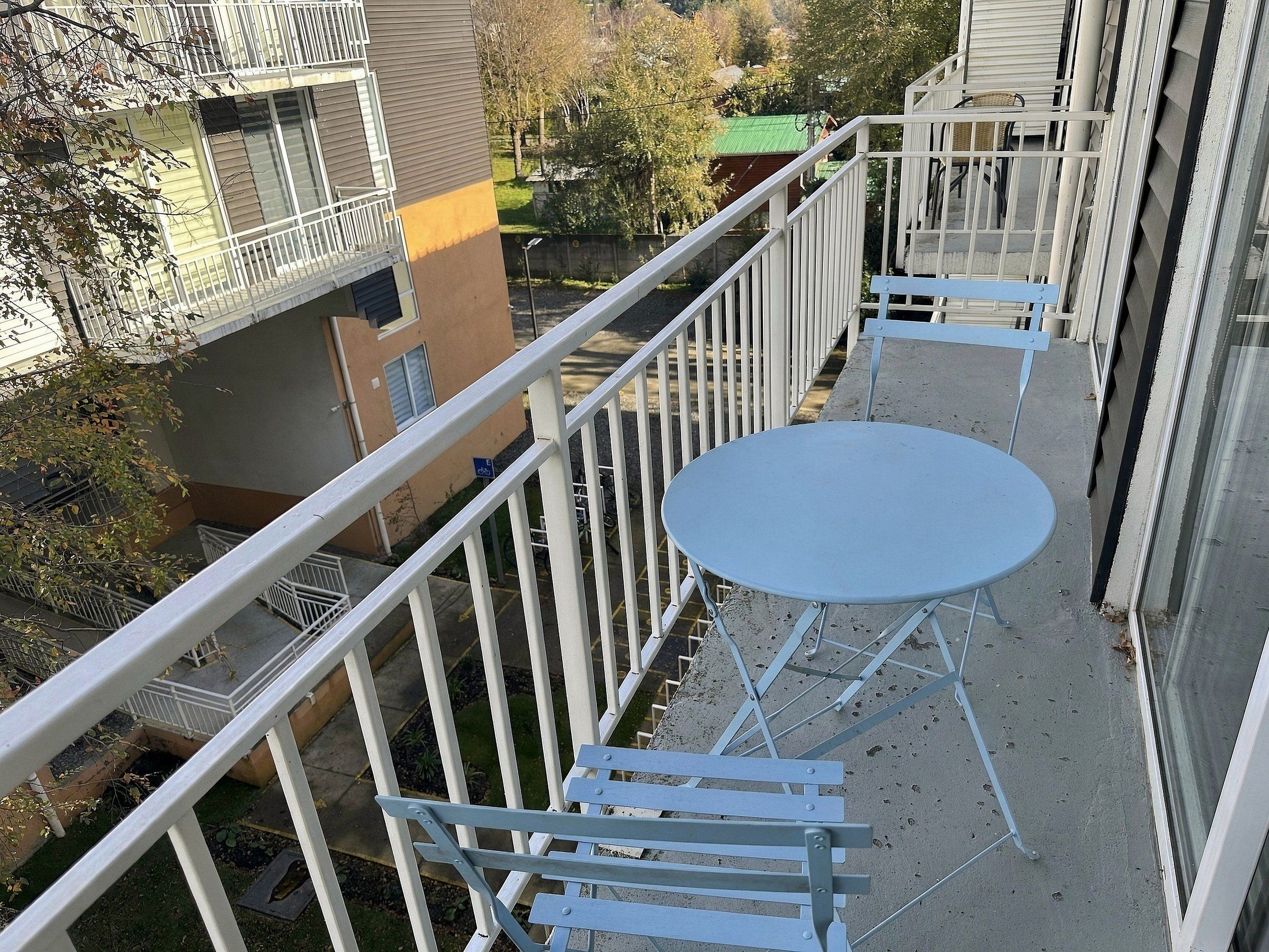 Un balcón con una mesa redonda y sillas, baranda de metal y vista de edificios cercanos.