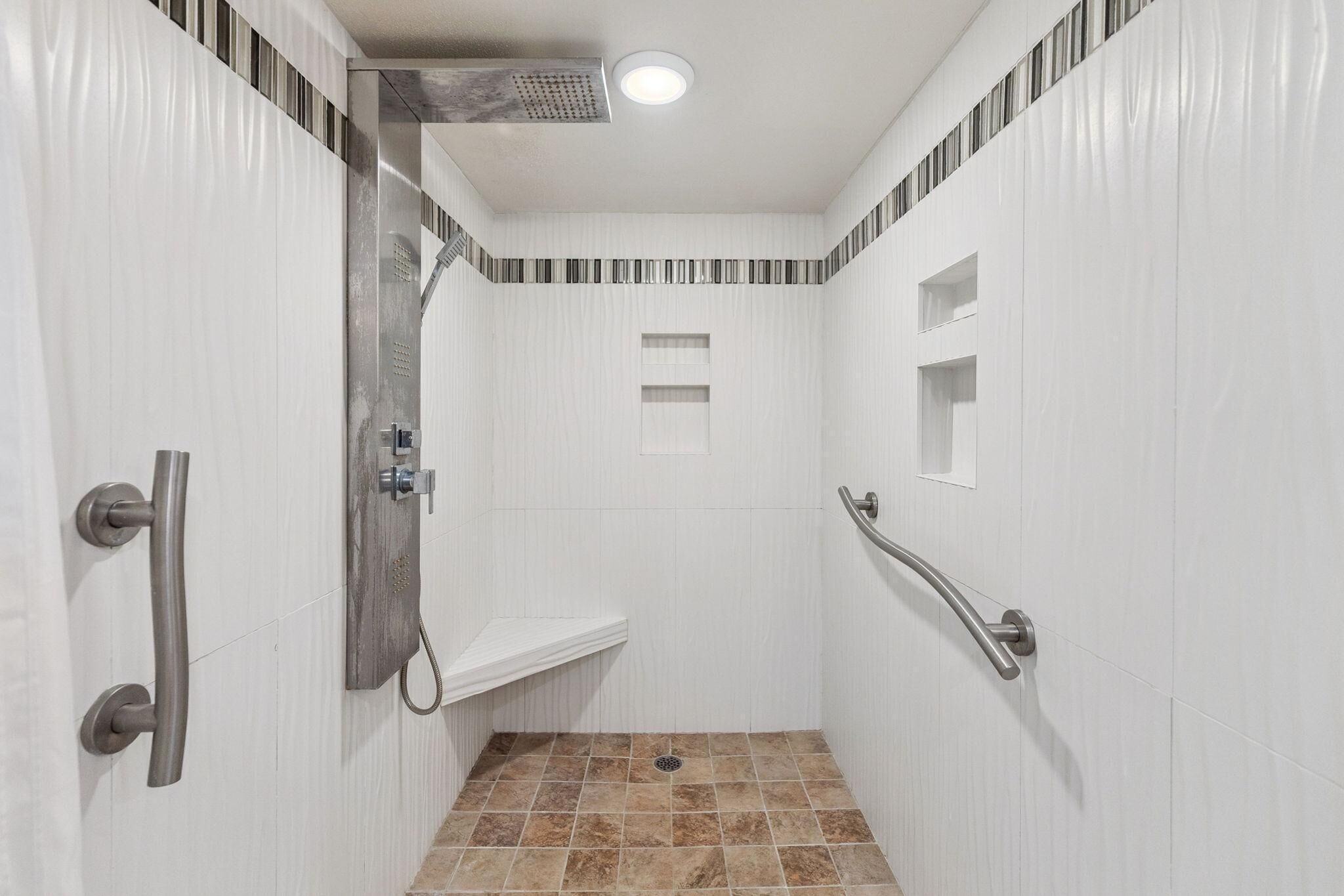 Un piccolo bagno rivestito di piastrelle bianche, con soffione, maniglione e ripiano.