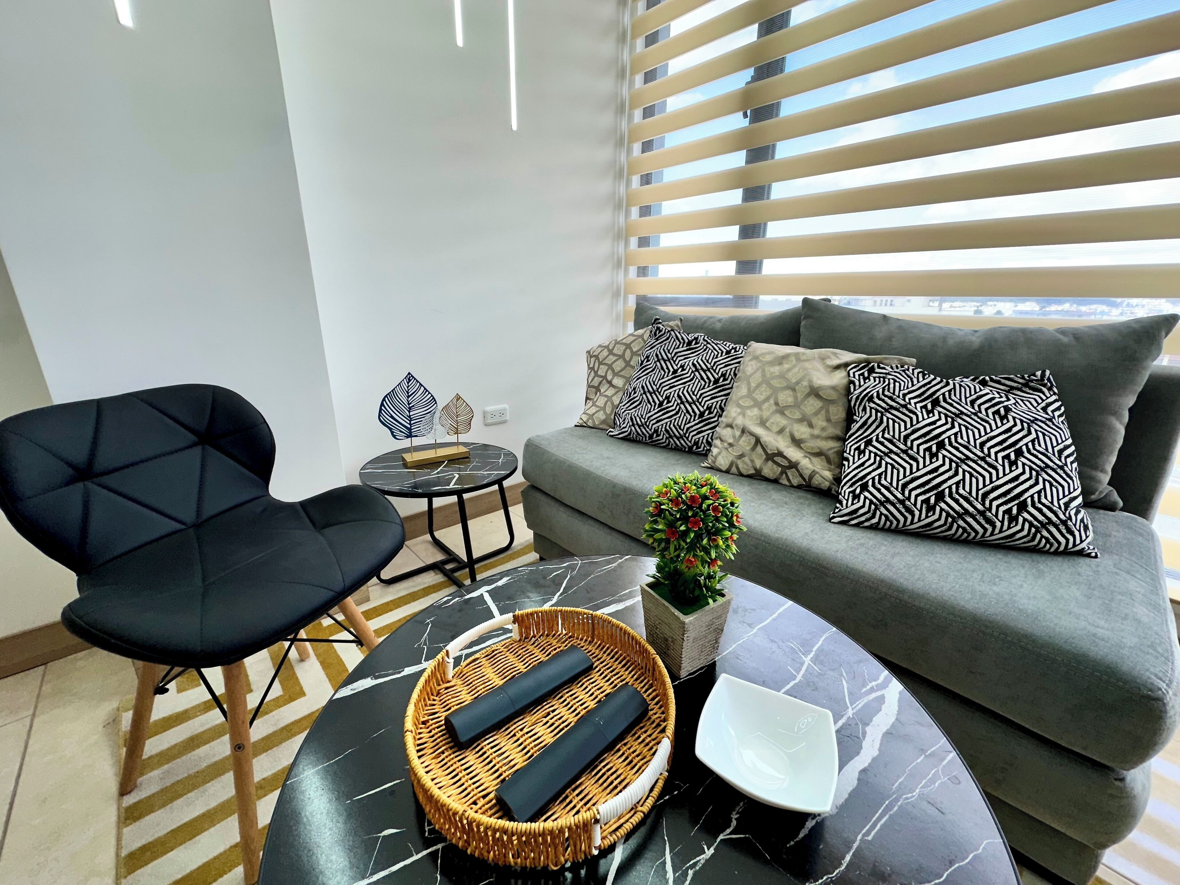 Una sala moderna con un sofá, un sillón negro, una mesa redonda y cojines decorativos.