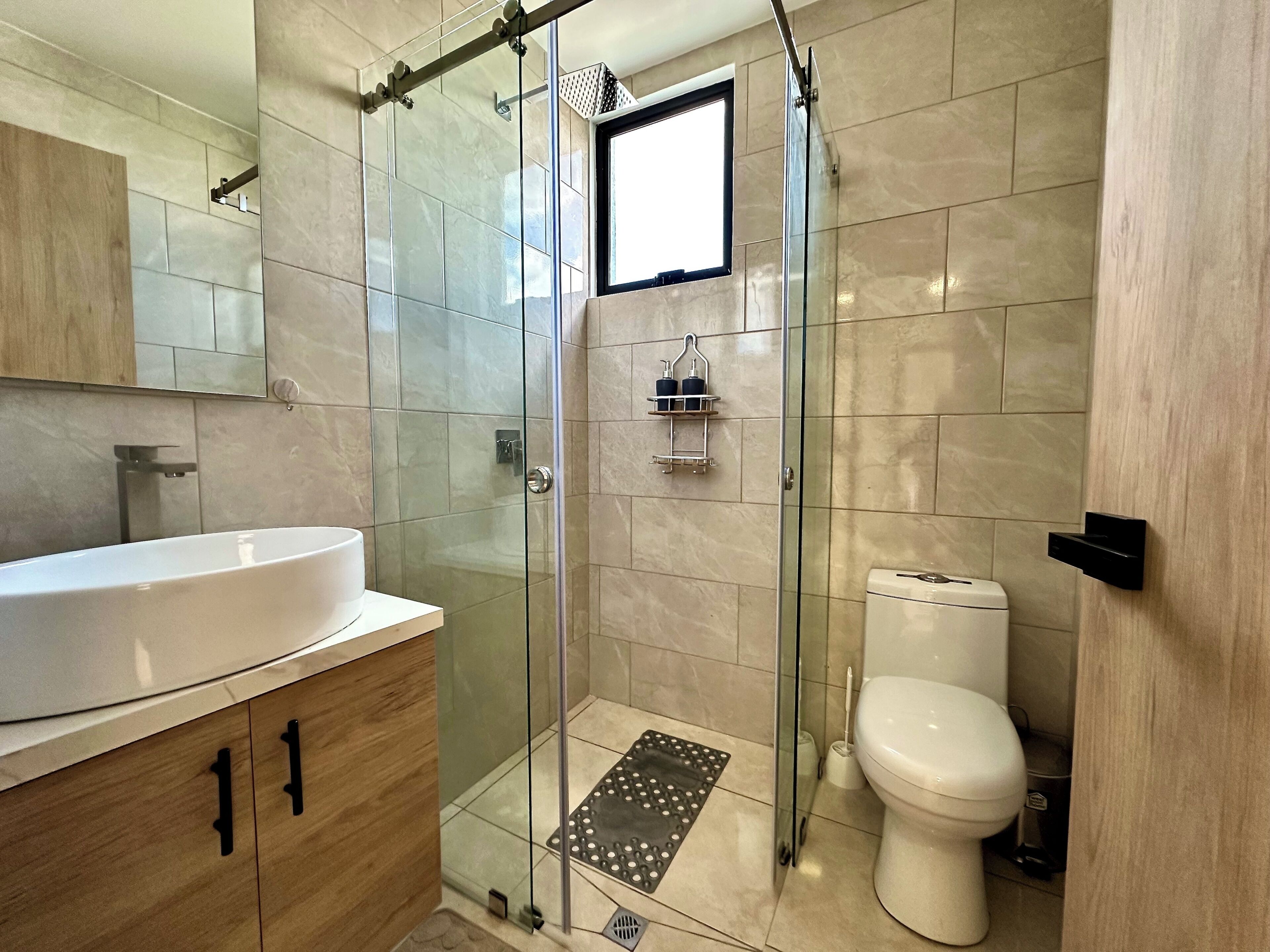 Un baño moderno con ducha envuelta en cristal, inodoro blanco, tocador con lavamanos y espejo.