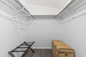 A small, white-walled room with a slanted ceiling, a metal rack, a brown suitcase, and a carpeted floor.