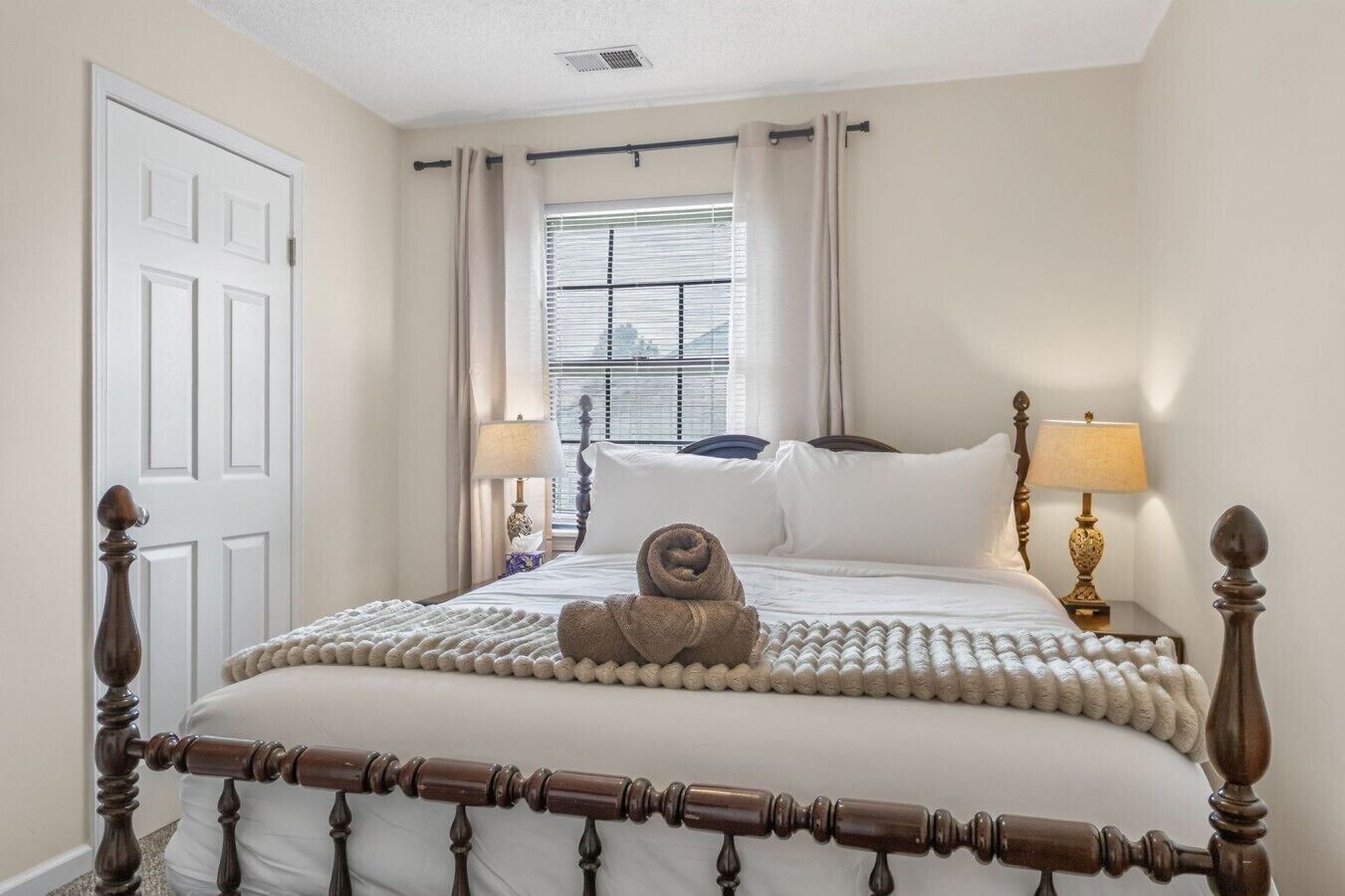 A bedroom with a wooden bed frame, white bedding, a window with blinds, and two bedside lamps.