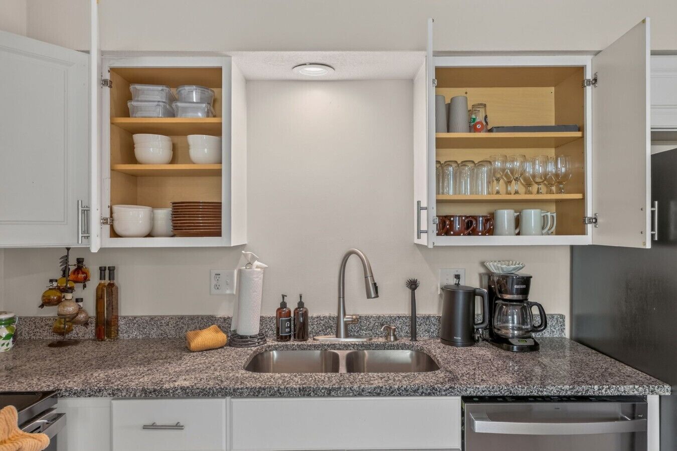 A kitchen with granite countertops, a stainless steel sink, and open cabinets with dishes and glassware.