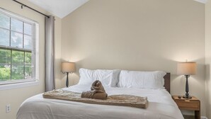 A neatly made bed with white linens, two bedside lamps, and a window with blinds.