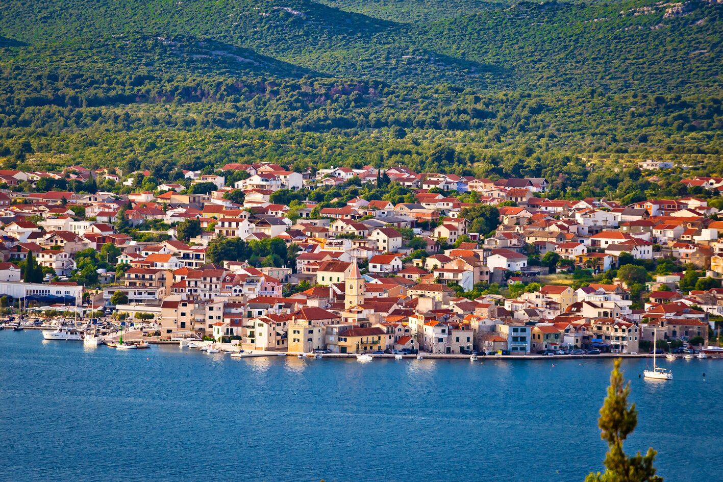 A coastal town with buildings densely packed along the waterfront, a marina with boats, and a backdrop of green hills.