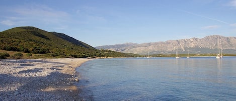 A pebble beach with boats, clear water, and mountains in the background.
