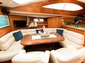 Interior de barco com mesa de madeira, assentos de couro branco, televisão, uma pequena réplica de veleiro e uma janela.