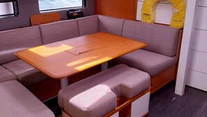 A compact boat interior with a wooden table, cushioned seating, a small shelf with items, and a window with a view of the sea. - Sun-Kissed Sailing Adventure in the Sporades Islands from Volos, Thessaly (Volos)