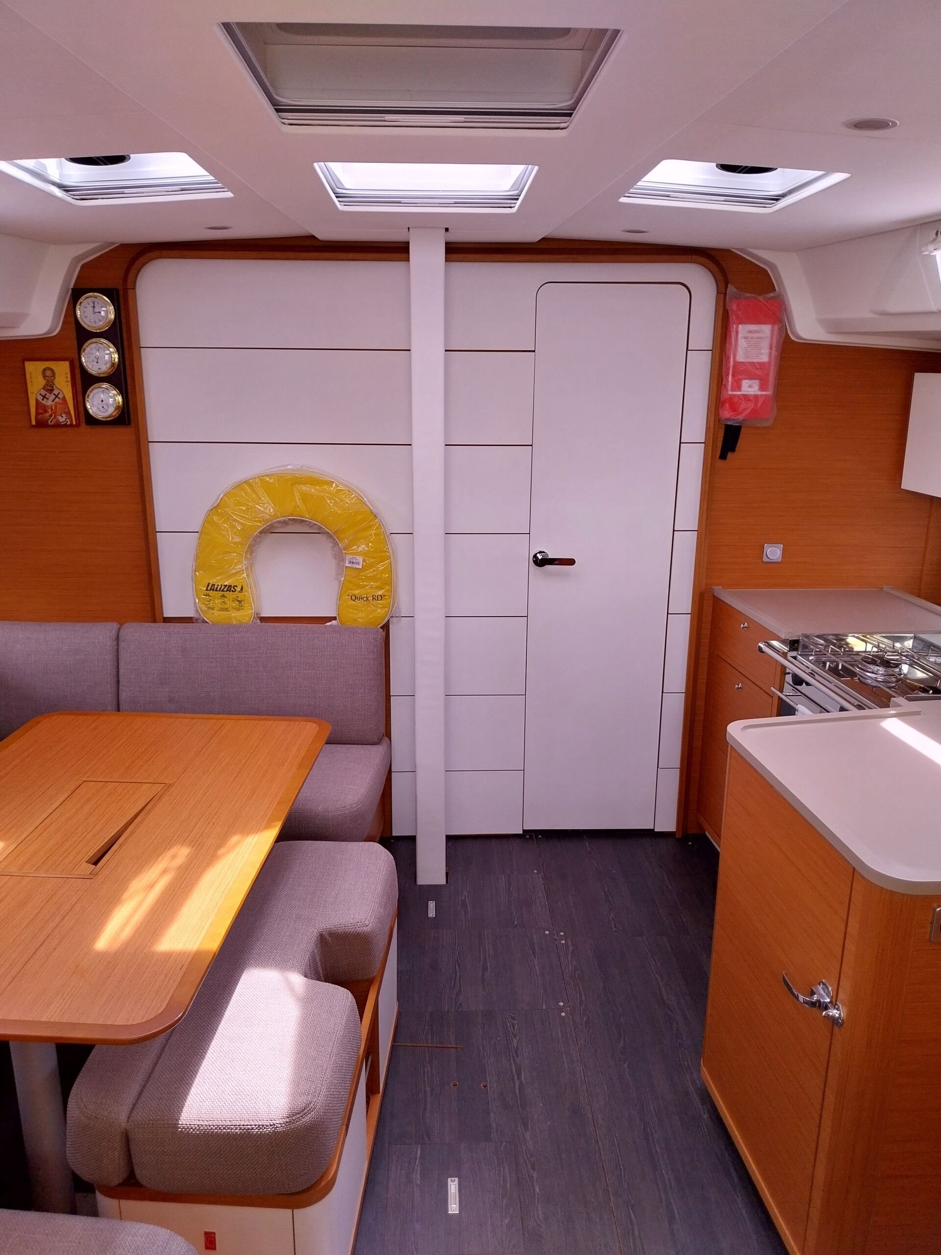 A compact interior with a dining table, cushioned seating, a kitchen area, and a door with a circular object on the wall.