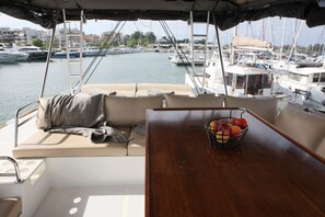 A boat's interior with a cushioned seating area, a wooden table with a fruit basket, and a view of a marina with boats. - Sun-kissed Sailing Adventure in the Ionian Islands (Preveza)