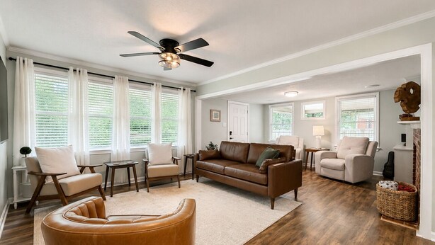 A spacious living room with a brown leather sofa, a brown armchair, and a coffee table. There is a ceiling fan, a fireplace, and a large window with blinds.