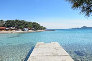 A clear waterfront with a concrete dock, a beach with buildings, and a forested hill in the background. - Sun-Kissed Sailing Adventure in Zadar, Croatia (Biograd na moru)