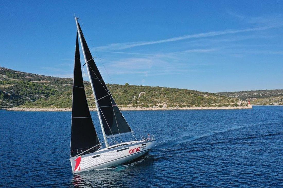 A sailboat with a red and white logo on the stern, sailing on a calm sea with a hilly coastline in the background.