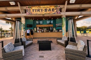 An outdoor tiki bar with wicker furniture, a bar counter, and a sign reading 'The Oasis TIKI BAR'.