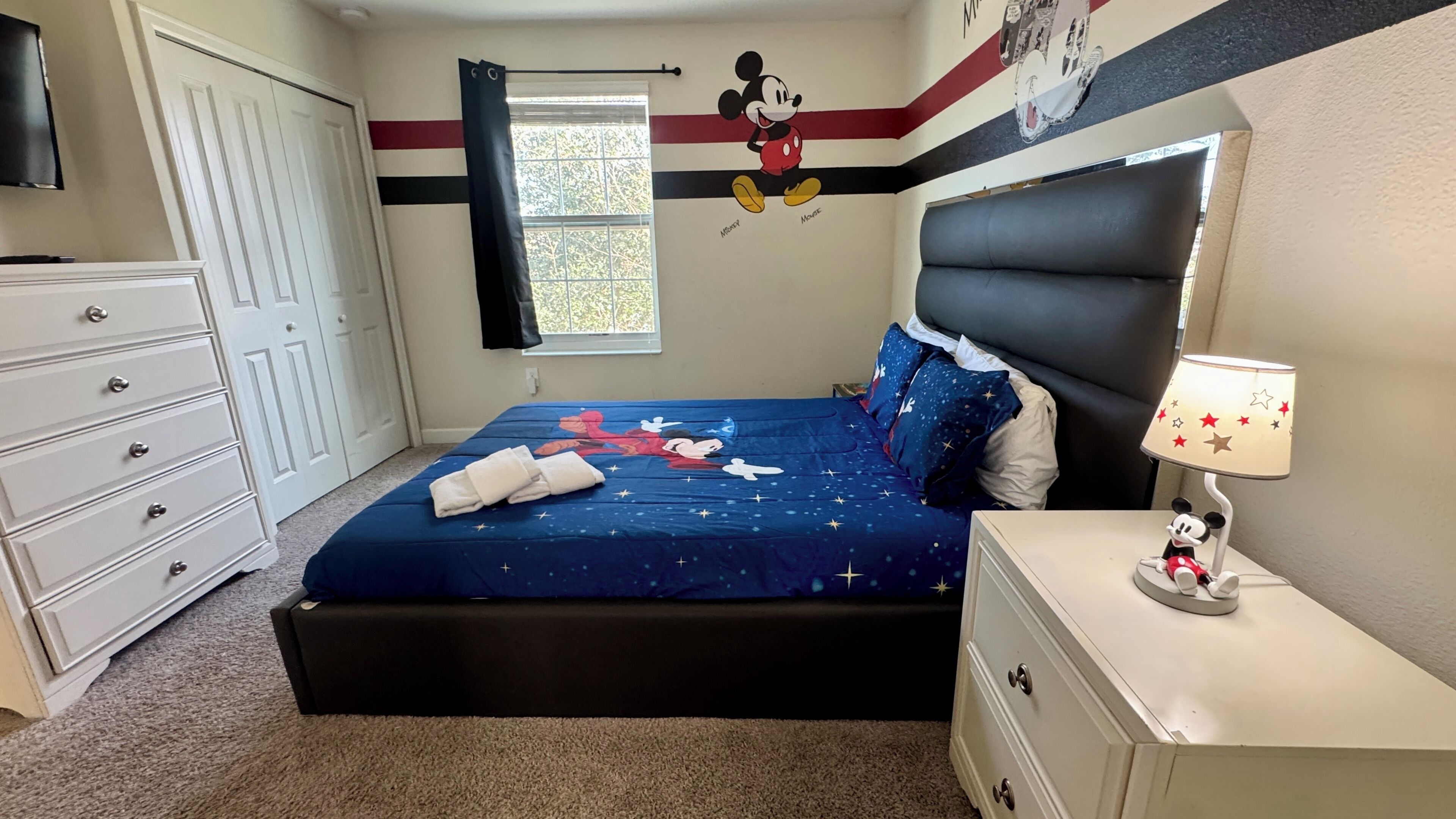 A bedroom with a bed, dresser, nightstand, and a Mickey Mouse wall decal.