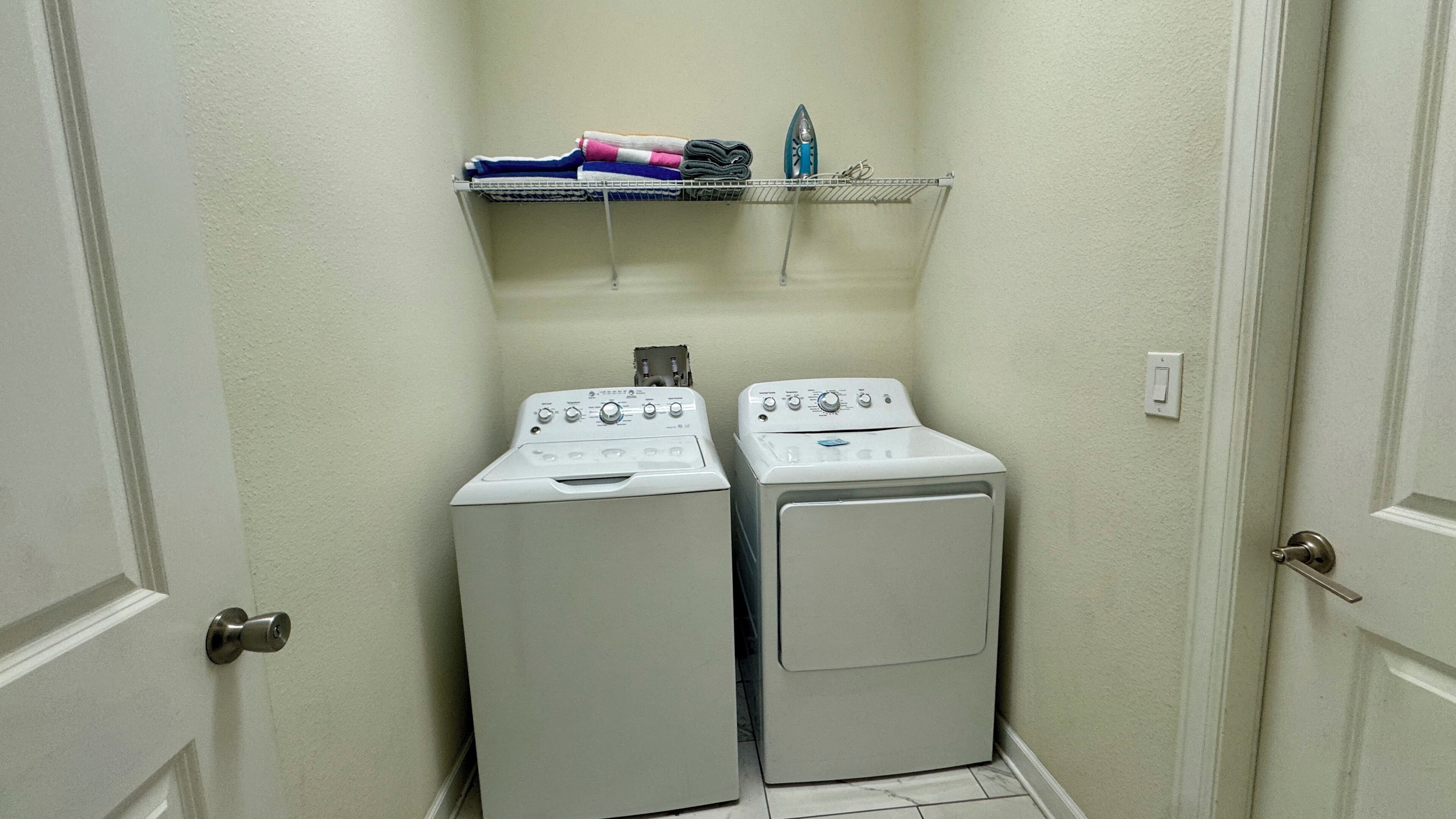 Een wasserij met twee gestapelde wasmachines en drogers, een plank met opgevouwen kleding, en een strijkijzer aan de muur.