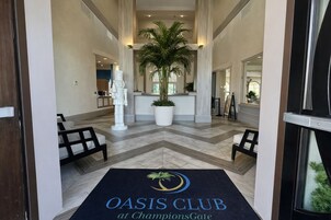 A hotel lobby with a reception desk, a large potted plant, and a doormat with 'OASIS CLUB at ChampionsGate' text.