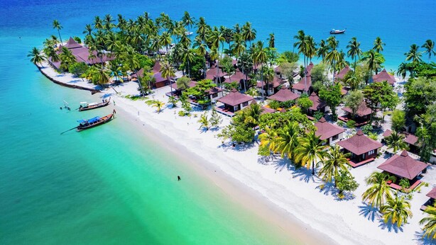 Vue aérienne d’un complexe hôtelier tropical doté de bungalows sur pilotis, d’une plage et d’eaux turquoise cristallines.