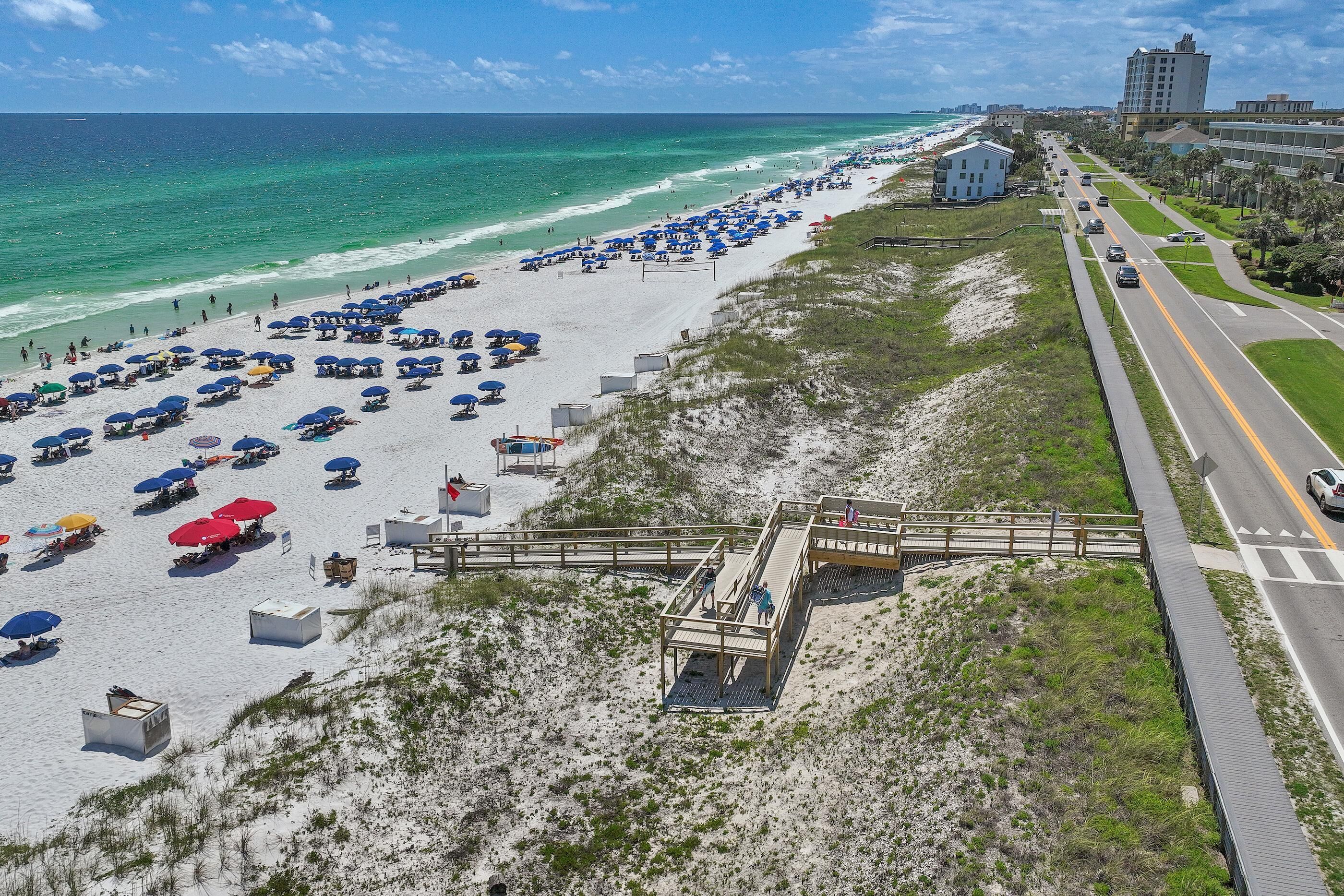 A beach with rows of umbrellas and beach chairs, a wooden boardwalk, and a road parallel to the beach.