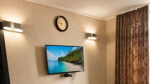 Ein modernes Hotelzimmer mit Wand-Fernseher, einer Holzkommode mit Pflanze, einem Esstisch mit Stühlen und einem großen Fenster mit Vorhängen.
