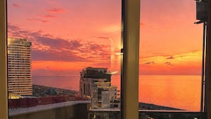 Ein Balkon mit Blick auf einen Sonnenuntergang über dem Meer, Gebäude im Hintergrund und ein Glastürrahmen.