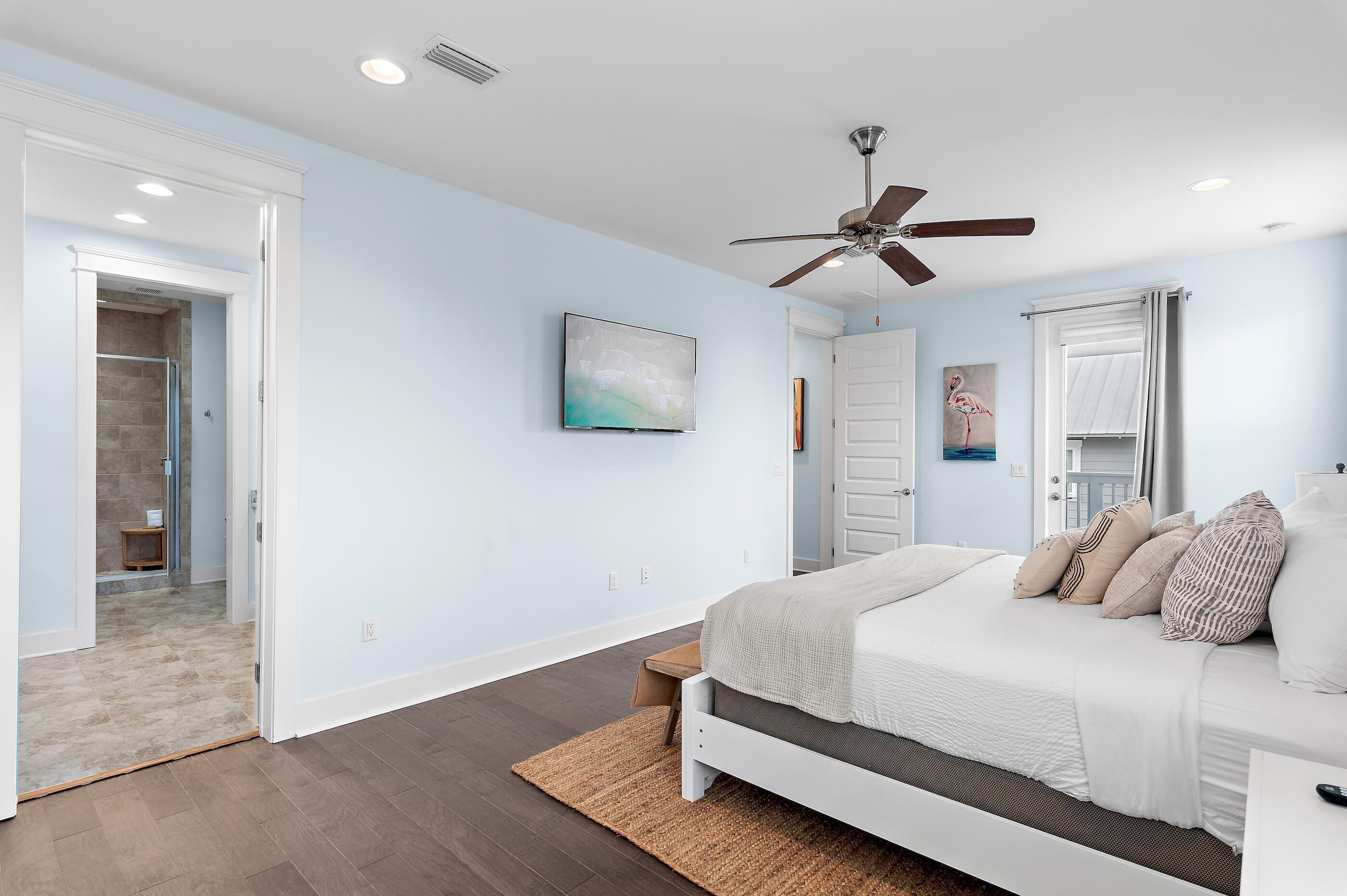A bedroom with a bed, a ceiling fan, a painting, and a door leading to a bathroom.