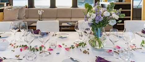 A dining area with a table set for a meal, a vase with flowers, and a view of the outdoors through large windows.