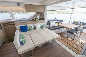 A boat interior with a sofa, table, and chairs, a small kitchen area, and a view of the water.