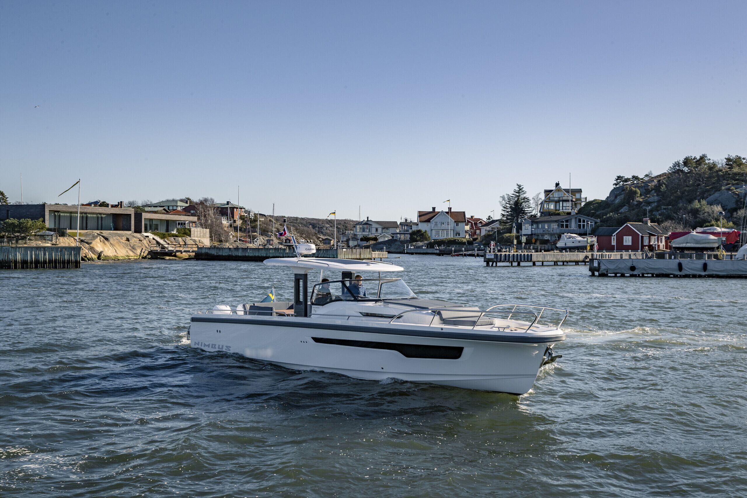 A white boat named 'NIMBUS' on calm water with a docked boat in the background, houses, and a hilly landscape.