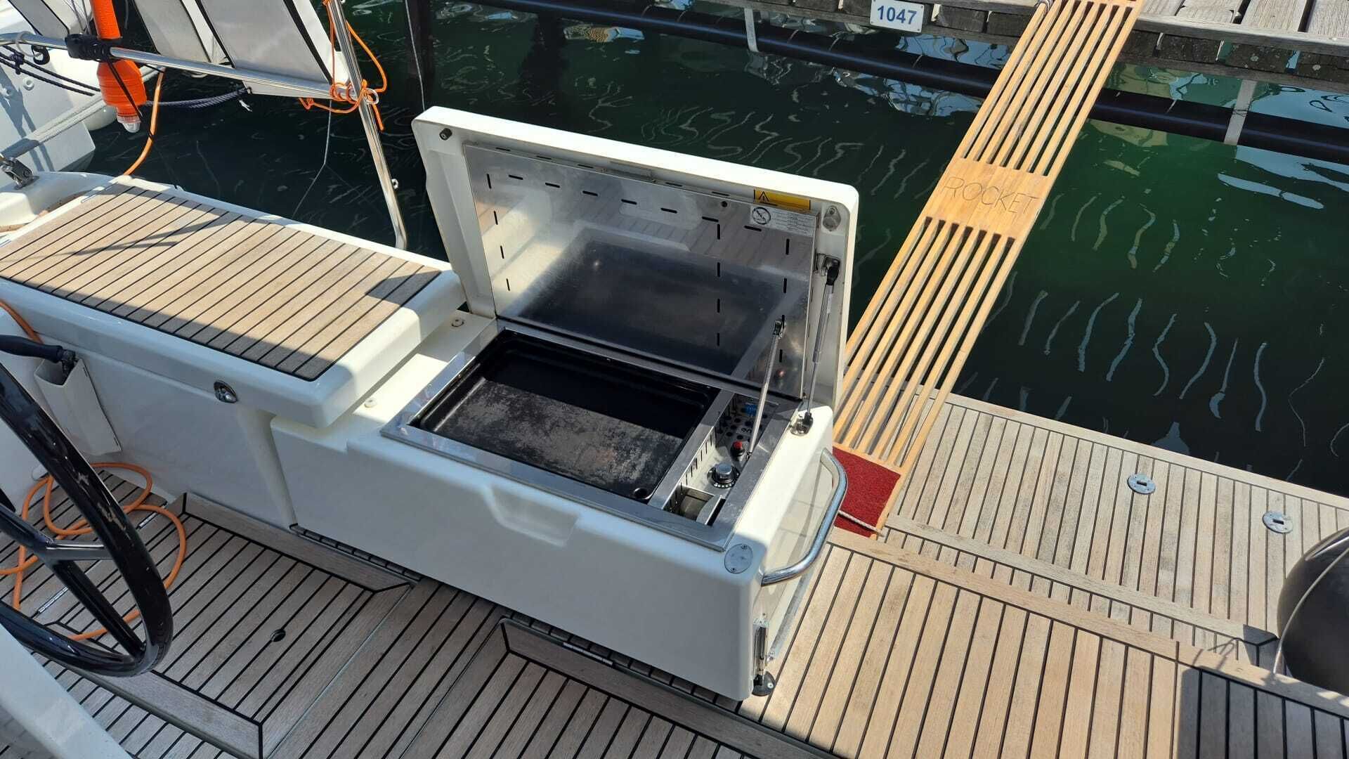 A boat's deck with a white, openable compartment, a wooden deck, and a water body in the background.