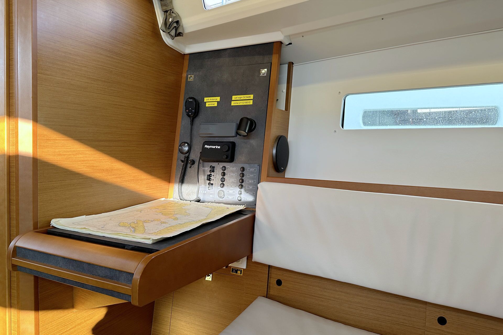 A compact cabin with a fold-down bed, a small table, a map, a Raymarine navigation device, and a window with a view.