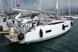 Eine weiße Segelyacht liegt in einer Marina festgemacht, mit mehreren Masten, einer deutschen Flagge und einem Schild mit der Aufschrift 'Perfect Charter'.