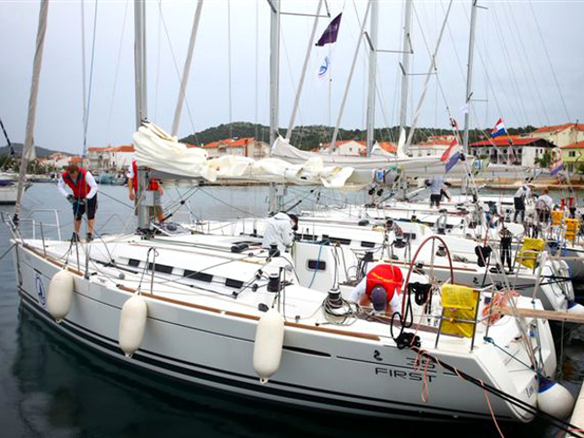 Un port de plaisance où sont amarrés plusieurs voiliers, dont certains arborent des drapeaux, et où une personne se tient sur un bateau.