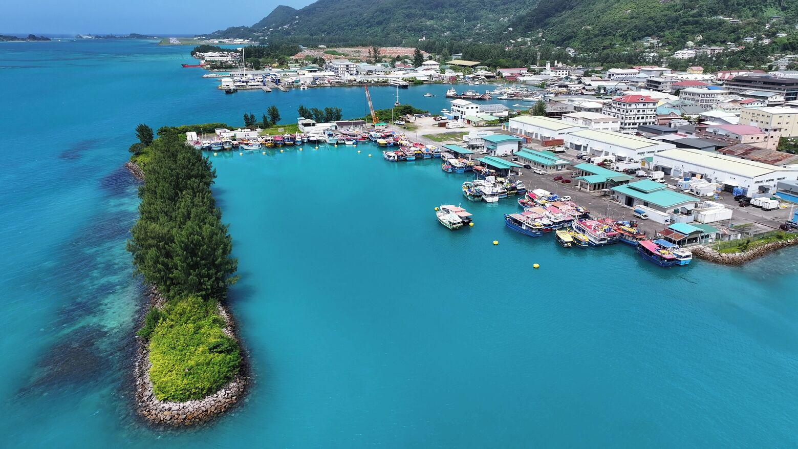 Vista aérea de un pueblo costero con un puerto deportivo, edificios y una pequeña isla con vegetación.