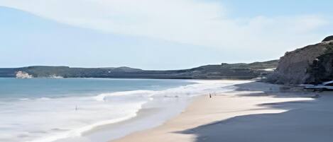 A beach with a person walking, clear skies, and distant hills.