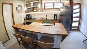 Ett kök med en köksö i mitten, barstolar, ett kylskåp och ett fönster med persienner.