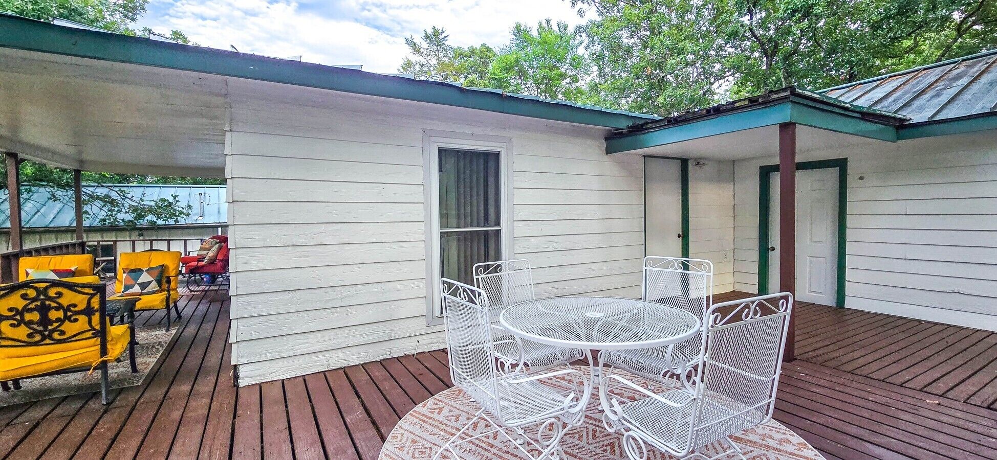 Une terrasse couverte avec une table et des chaises en métal blanc, une terrasse en bois et un petit bâtiment doté d’un toit végétalisé.