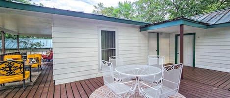 A covered patio with a white metal table and chairs, a wooden deck, and a small building with a green roof.